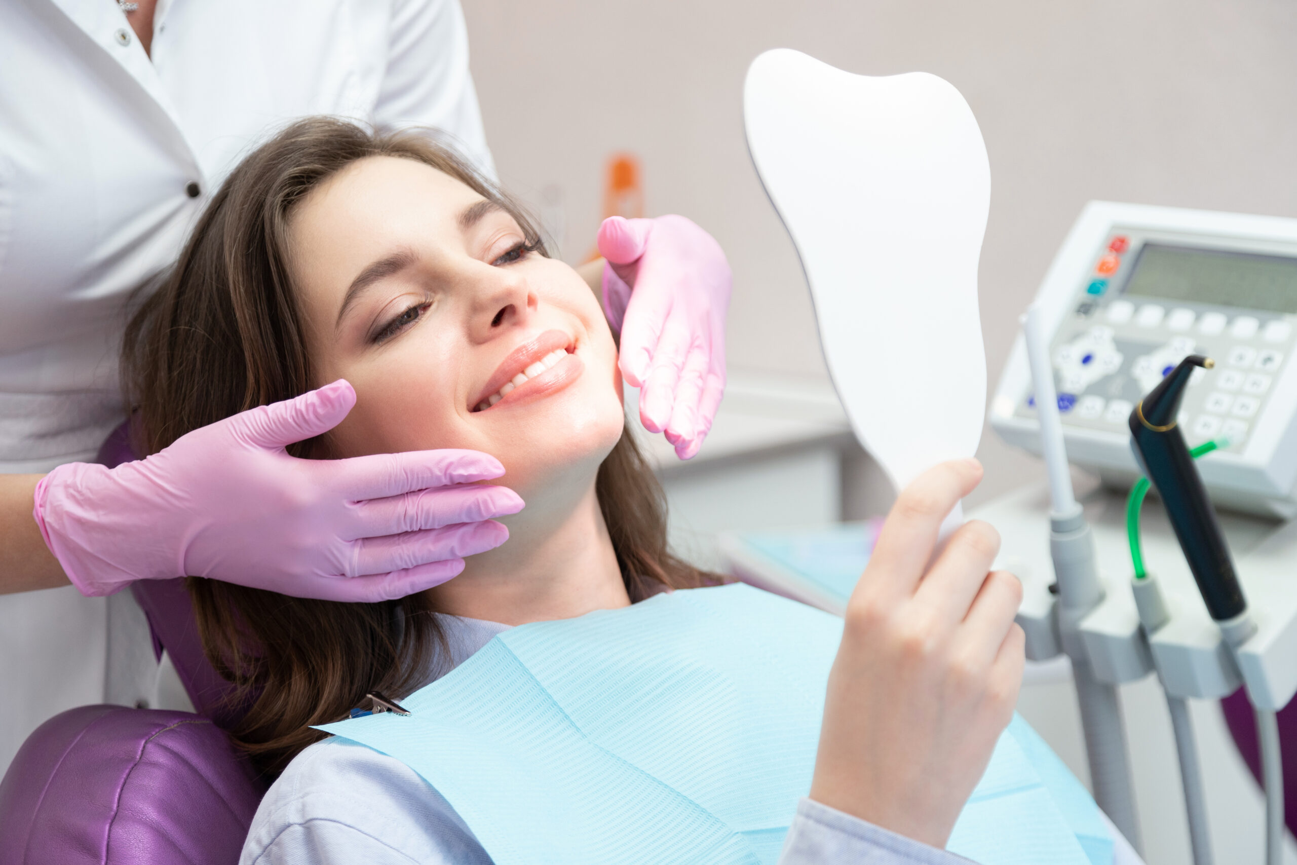 Young glad smiling woman patient looking at mirror after teeth whitening procedure, caries treatment, toothache elimination. Pretty happy girl at stomatologist appointment in dental clinic